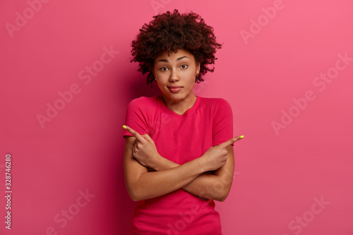 Questioned dark skinned woman makes choice, shows two ways, points sideways, has hesitant expression, wears casual t shirt, poses over rosy background, looks with bewilderment or puzzlement.