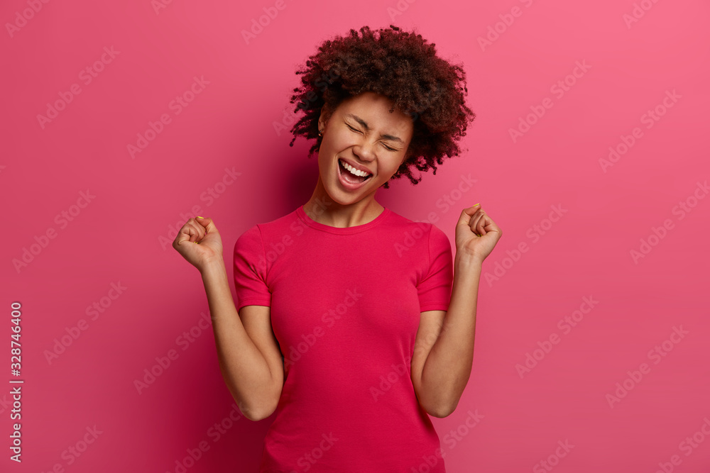 People, emotions, triumph and success concept. Happy Afro American teenage girl celebrates victory, raises fists up, has upbeat mood, dressed in casual clothing, isolated on pink background. People, emotions, triumph and success concept. Happy Afro American teenage girl celebrates victory, raises fists up, has upbeat mood, dressed in casual clothing, isolated on pink background.