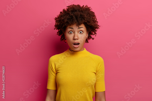 Fascinated concerned woman with Afro hair gasps from fear, faces difficulties, stands troubled, wears yellow casual t shirt, being in troublesome situation, isolated over pink wall, keeps mouth opened © wayhome.studio 