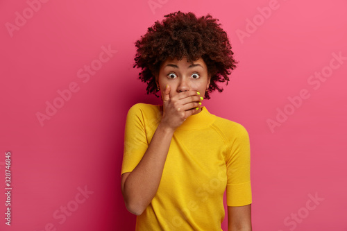 Scared dark skinned woman covers mouth, holds breath from fright, stands speechless, finds out shocking truth or news, dressed in casual yellow clothes, isolated on pink background, looks worried © Wayhome Studio