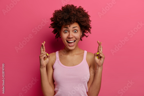 Beautiful smiling dark skinned woman wishes good luck, believes dreams or wish come true, laughs happily, stands with crossed fingers, wears casual clothes, isolated over pink bright background