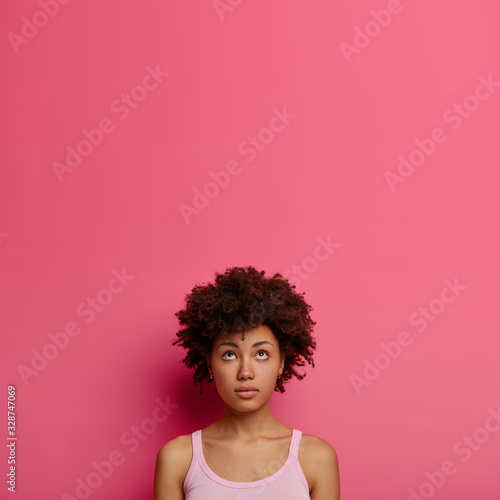 Natural curly African American woman looks thoughtfully above, has some thought in mind, wears casual clothes, stands against pink background, copy space for your promotion, recalls something