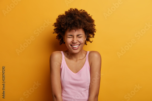 Portrait of positive ethnic woman squints face, smiles happily, shows white teeth, being in high spirit, enjoys day off, listens good jokes from friend, wears casual vest, models against yellow wall