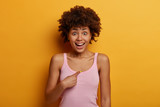 Pleased dark skinned model asks who me, indicates at herself, has surprised glad expression, giggles happily, has widely opened eyes, isolated over yellow background. Am I appropriate candidate?