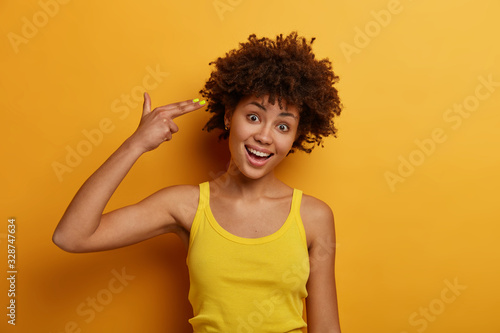 Funny joyful dark skinned woman commits suicide, makes finger gun pistol, tilts head and shoots in temple, foolishes around and feels bored, wears yellow casual clothes. People, lifestyle, emotions