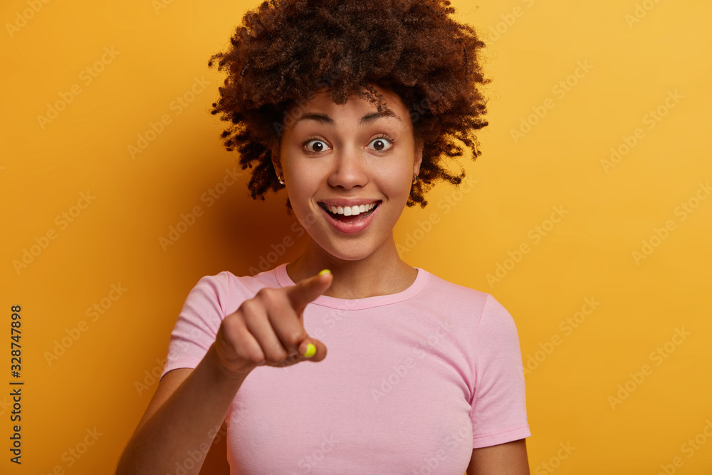 Cropped image of pretty joyful woman with toothy smile points directly at camera, sees something amazing in front, wears t shirt, has curious happy expression, isolated on yellow background. Cropped image of pretty joyful woman with toothy smile points directly at camera, sees something amazing in front, wears t shirt, has curious happy expression, isolated on yellow background.