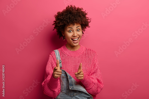 Only you. Glad curly haired Afro American woman points straightly at camera with finger gun gesture or hand gun, has glad expression, dressed in loose knitted sweater, advertises product and sale