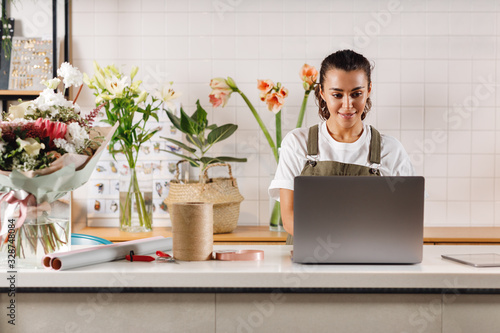 Young flower shop owner using laptop at counter. Woman florist standing at table. 