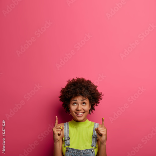 Look there. Pretty satisfied woman has happy expression  promots goods  indicates above  says your promotion here  demonstrates copy space on pink wall for your advertising content. Spring sale