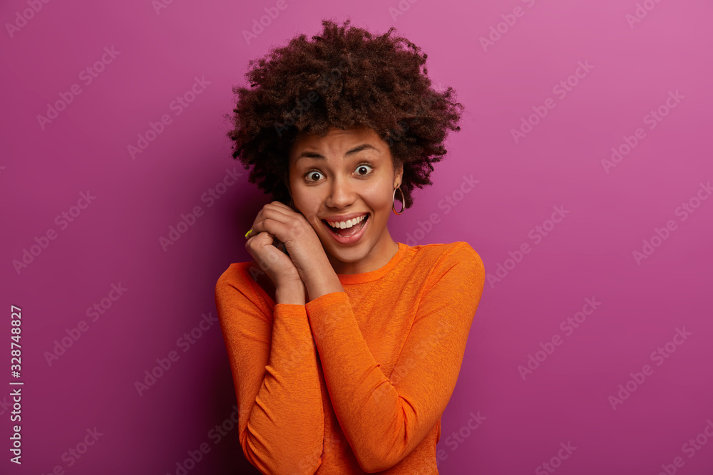 Isolated shot of pretty ethnic young woman keeps hands near face, smiles pleasantly, being in good mood, dressed in casual orange jumper, has natural curly hair, poses against purple background Isolated shot of pretty ethnic young woman keeps hands near face, smiles pleasantly, being in good mood, dressed in casual orange jumper, has natural curly hair, poses against purple background