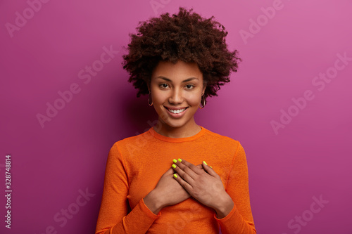Pleased girl in orange jumper presses palms to heart, makes thankful gesture, touched with cordial congratulations, smiles positively, isolated over purple background. Acknowledgement concept