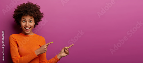 Happy reaction on advert. Positive excited woman has Afro hairstyle, dressed in orange jumper, has widely opened eyes, shows blank space on purple background for your promotion or information photo