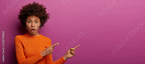 Portrait of emotional woman stares with surprise, shocked by decreased prices, indicates right on copy space, has astonished expression, demonstrates something amazing, dressed in bright clothes
