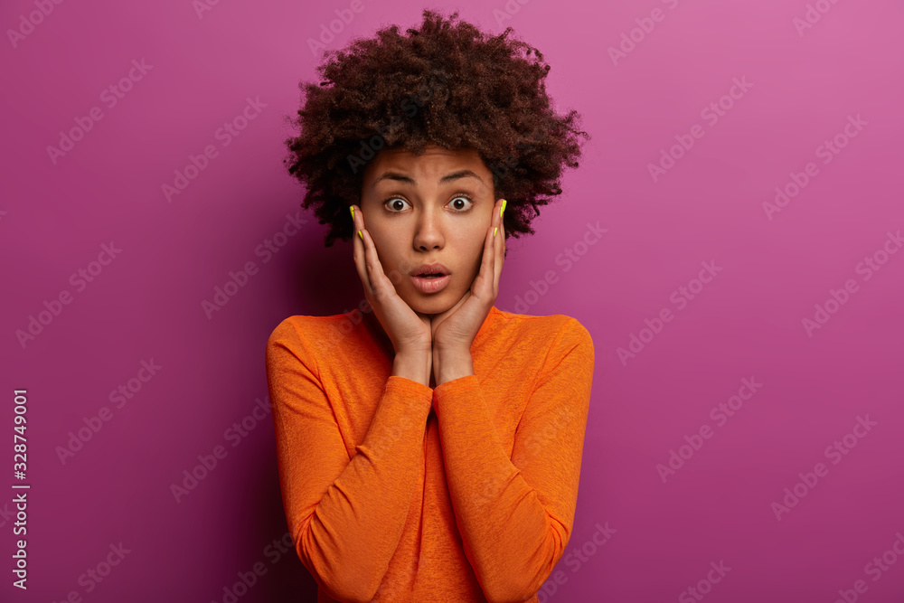 Fascinated Afro American woman grabs cheeks, dressed in orange jumper, realizes she forgot about something important, receives big fine, poses against purple background. Human face expressions Fascinated Afro American woman grabs cheeks, dressed in orange jumper, realizes she forgot about something important, receives big fine, poses against purple background. Human face expressions