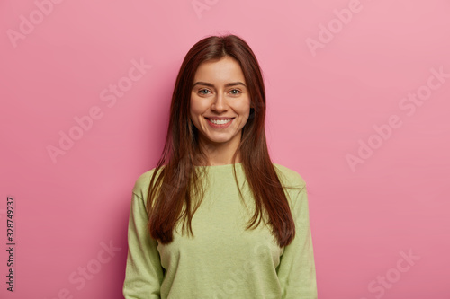 Portrait of attractive woman has healthy skin  has toothy smile  looks directly at camera  wears green jumper  has long straight hair  poses against pink pastel background. Face expressions concept