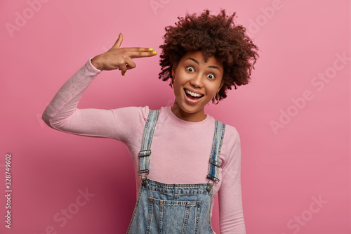 Positive carefree woman shoots with finger gun gesture at temple, commits suicide, foolishes around, looks joyfully at camera, wears denim overalls and poloneck, isolated on pink pastel background