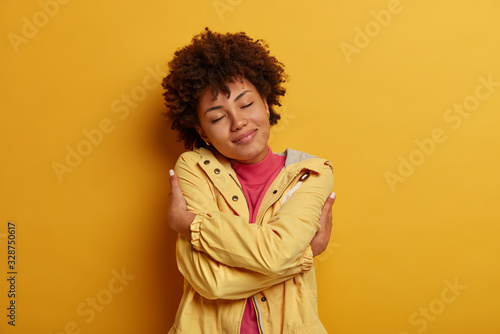 Selfishness and self love concept. Portrait of pleased dark skinned curly female model hugs herself, crosses hands over body, keeps eyes closed, wears jacket, poses against yellow background © wayhome.studio 