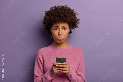 Fotografie, Obraz Unhappy disappointed woman with Afro hair, purses lower lip, holds smartphone, s