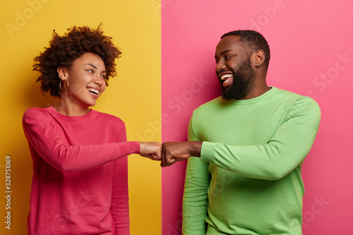 Positive dark skinned young woman and man bump fists, agree to be one team, look happily at each other, celebrates completed task, wear pink and green clothes, pose indoor, have successful deal photo