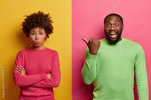 Happy bearded man points thumb on sad disappointed woman who feels offended and insulted, disagrees with someones opinion. Ethnic couple expess different emotions, pose indoor over two colored wall photo