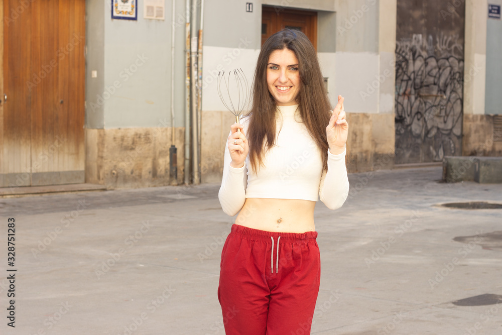Portrait of attractive caucasian young woman holding neural massager, keeping finger crossed, in the city, white top and red pants, dark air. Place for your text in copy space. Portrait of attractive caucasian young woman holding neural massager, keeping finger crossed, in the city, white top and red pants, dark air. Place for your text in copy space.