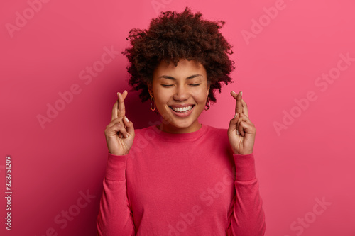 Headshot of pretty curly superstitious woman crosses fingers, prays for better, smiles gently, closes eyes, wears pink jumper, poses indoor over bright background, anticipates important results