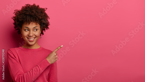 Horizontal shot of pretty woman with curly hairstyle, has tender smile, indicates at upper right corner, shows copy space for your advertising content, wears pink clothes. People and promotion concept