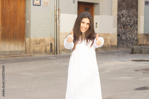 Portrait of happy confidence caucasian young woman showing thumbs up gesture. Doctor, nurse, pharmacist, beauty therapist, scientist, healthcare, in the city, in white coat uniform, dark air. Place fo