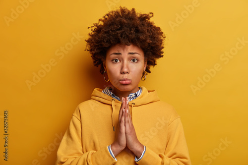 Let me please. Sad pleading African American woman asks for permission, holds hands in pray, says forgive me, poses against yellow background, wears sweatshirt. Begging and saying forgive me. photo