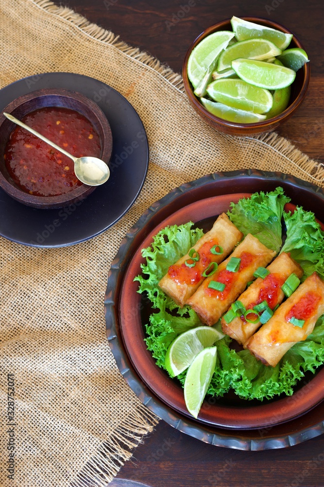 Portrait of spring rolls on a bed of green lettuce next to a small bowl of sweet chilly sauce and a bowl of limes on a hessian background Portrait of spring rolls on a bed of green lettuce next to a small bowl of sweet chilly sauce and a bowl of limes on a hessian background