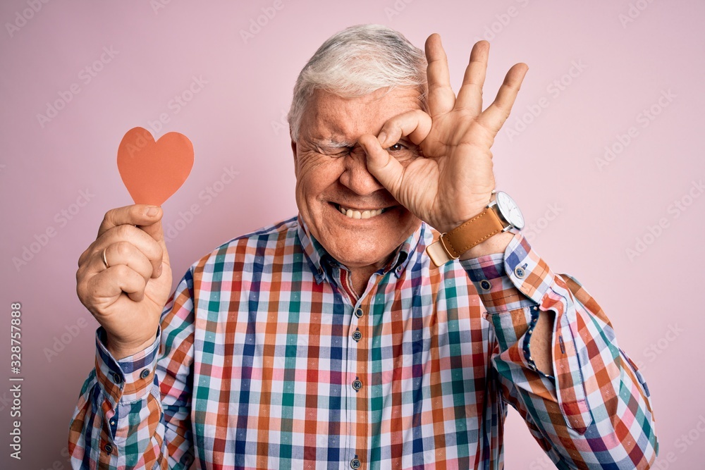 Naklejka premium Senior handsome hoary romantic man holding red paper heart shape over pink background with happy face smiling doing ok sign with hand on eye looking through fingers
