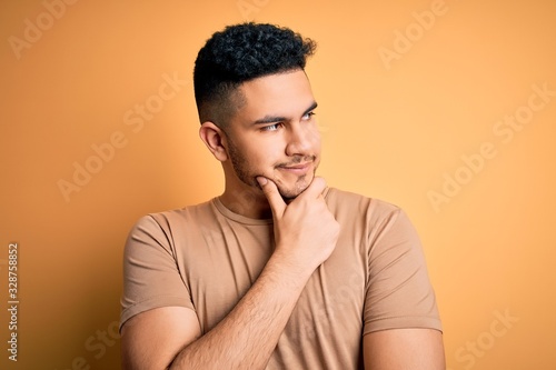 Young handsome man wearing casual t-shirt standing over isolated yellow background with hand on chin thinking about question, pensive expression. Smiling with thoughtful face. Doubt concept. © Krakenimages.com