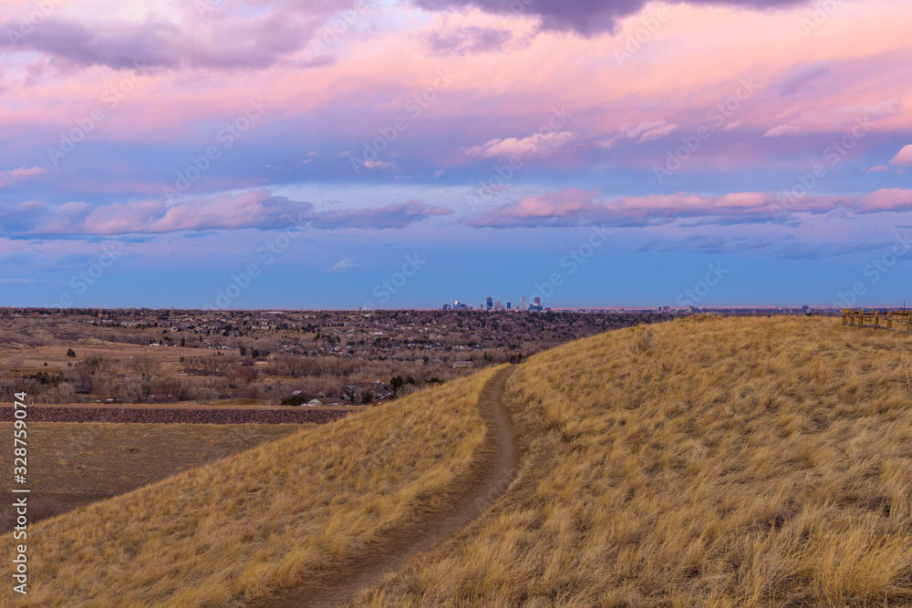 Fototapeta premium Sunset Hiking Trail - Colorful winter sunset view of a hiking trail winding at hillside, with Denver downtown skyline in background. Denver-Lakewood, Colorado, USA.