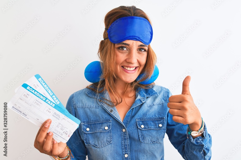 Young redhead tourist woman wearing sleeping mask and neck pillow showing boarding pass happy with big smile doing ok sign, thumb up with fingers, excellent sign