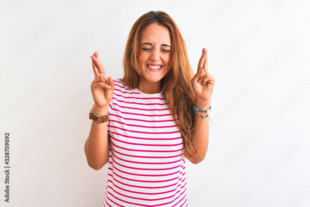 Young redhead woman wearing striped casual t-shirt stading over white isolated background gesturing finger crossed smiling with hope and eyes closed. Luck and superstitious concept. Young redhead woman wearing striped casual t-shirt stading over white isolated background gesturing finger crossed smiling with hope and eyes closed. Luck and superstitious concept.