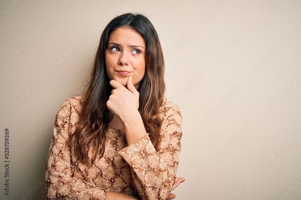 Young beautiful brunette woman wearing casual t-shirt standing over white background with hand on chin thinking about question, pensive expression. Smiling with thoughtful face. Doubt concept. Young beautiful brunette woman wearing casual t-shirt standing over white background with hand on chin thinking about question, pensive expression. Smiling with thoughtful face. Doubt concept.