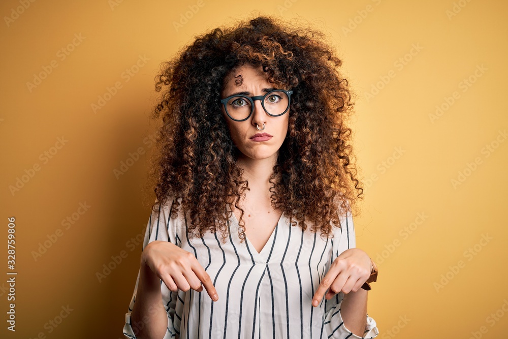Young beautiful woman with curly hair and piercing wearing striped shirt and glasses Pointing down looking sad and upset, indicating direction with fingers, unhappy and depressed. Young beautiful woman with curly hair and piercing wearing striped shirt and glasses Pointing down looking sad and upset, indicating direction with fingers, unhappy and depressed.
