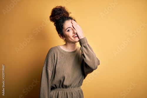 Young beautiful brunette woman with curly hair and piercing wearing casual dress covering one eye with hand, confident smile on face and surprise emotion.