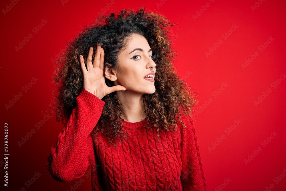 Young beautiful woman with curly hair and piercing wearing casual red sweater smiling with hand over ear listening an hearing to rumor or gossip. Deafness concept. Young beautiful woman with curly hair and piercing wearing casual red sweater smiling with hand over ear listening an hearing to rumor or gossip. Deafness concept.
