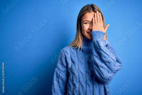 Young beautiful blonde woman wearing winter wool sweater over blue isolated background covering one eye with hand, confident smile on face and surprise emotion.
