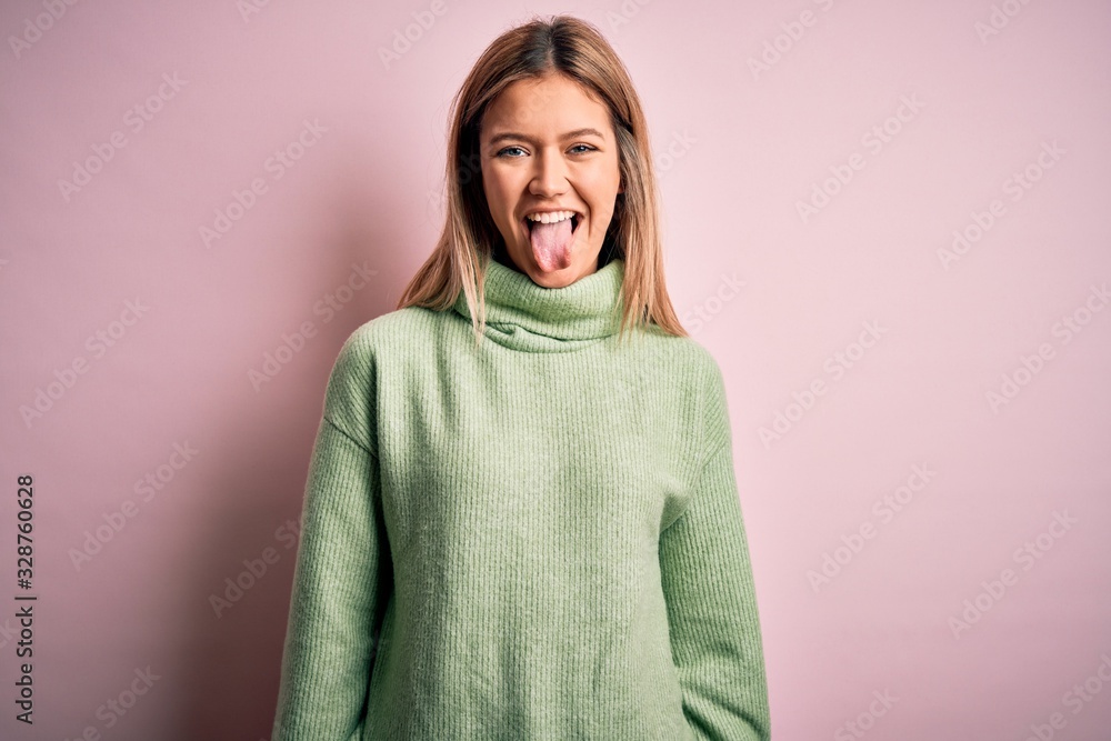 Young beautiful blonde woman wearing winter wool sweater over pink isolated background sticking tongue out happy with funny expression. Emotion concept. Young beautiful blonde woman wearing winter wool sweater over pink isolated background sticking tongue out happy with funny expression. Emotion concept.