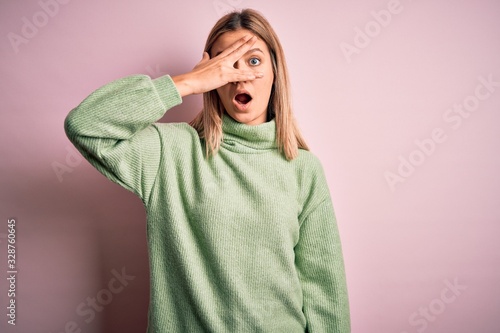 Young beautiful blonde woman wearing winter wool sweater over pink isolated background peeking in shock covering face and eyes with hand, looking through fingers with embarrassed expression. © Krakenimages.com