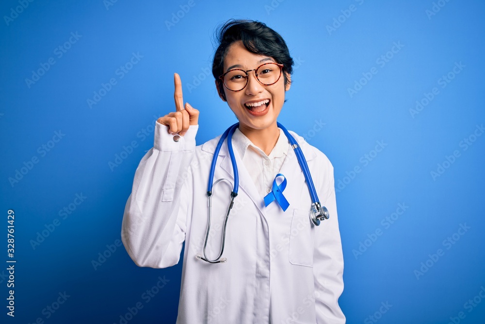 Young beautiful asian doctor girl wearing stethoscope and coat with blue cancer ribbon pointing finger up with successful idea. Exited and happy. Number one. Young beautiful asian doctor girl wearing stethoscope and coat with blue cancer ribbon pointing finger up with successful idea. Exited and happy. Number one.