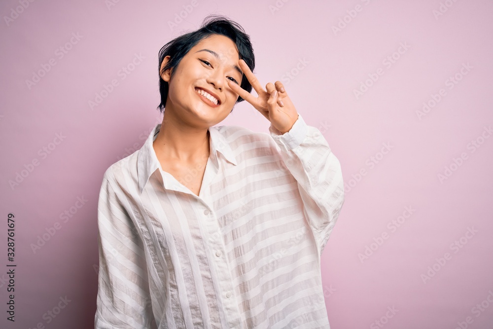 Young beautiful asian girl wearing casual shirt standing over isolated pink background Doing peace symbol with fingers over face, smiling cheerful showing victory