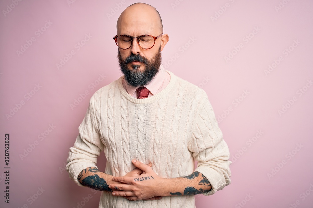Handsome bald man with beard and tattoo wearing glasses and sweater over pink background with hand on stomach because indigestion, painful illness feeling unwell. Ache concept. Handsome bald man with beard and tattoo wearing glasses and sweater over pink background with hand on stomach because indigestion, painful illness feeling unwell. Ache concept.