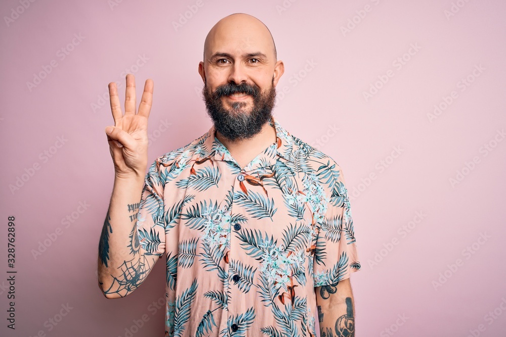 Naklejka premium Handsome bald man with beard and tattoo wearing casual floral shirt over pink background showing and pointing up with fingers number three while smiling confident and happy.