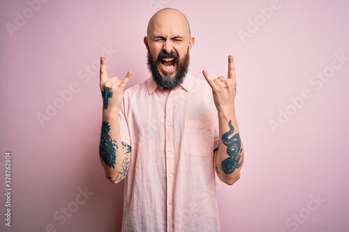 Handsome bald man with beard and tattoo wearing casual shirt over isolated pink background shouting with crazy expression doing rock symbol with hands up. Music star. Heavy concept. photo