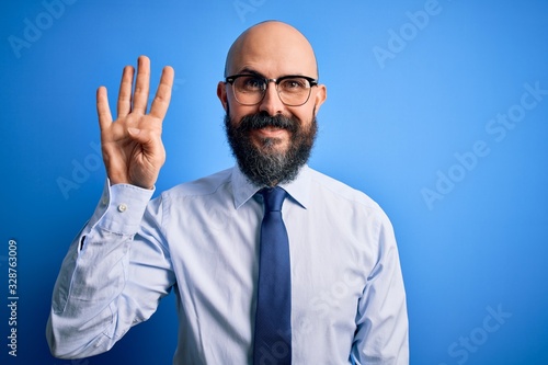 Handsome business bald man with beard wearing elegant tie and glasses over blue background showing and pointing up with fingers number four while smiling confident and happy.