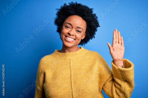 Young beautiful African American afro woman with curly hair wearing yellow casual sweater Waiving saying hello happy and smiling, friendly welcome gesture photo
