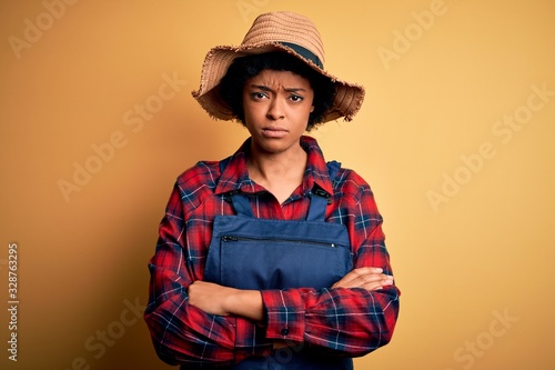 Young African American afro farmer woman with curly hair wearing apron and hat skeptic and nervous, disapproving expression on face with crossed arms. Negative person.